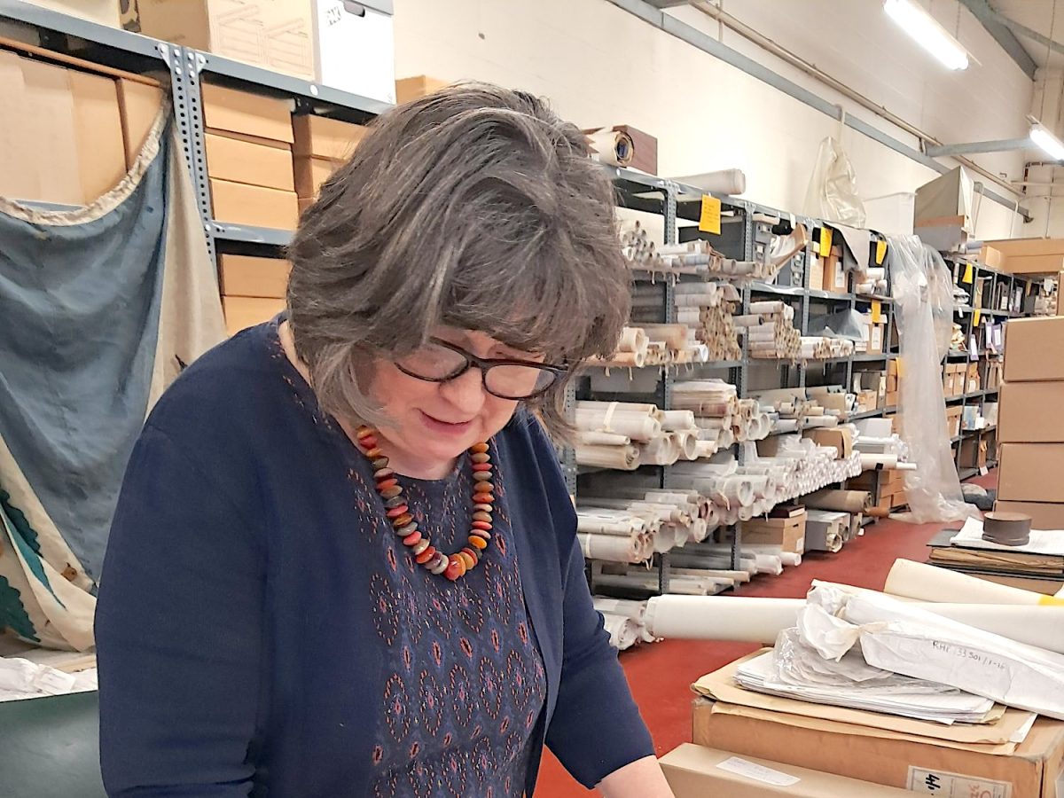 A photo of a women, in the Ballast Trust organising archival materials, behind her there are shelves with boxes and rolled up sheets of paper.